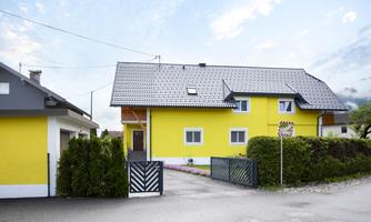A yellow house with a gray roof and a beautiful garden. The entrance is accessible through a black gate.