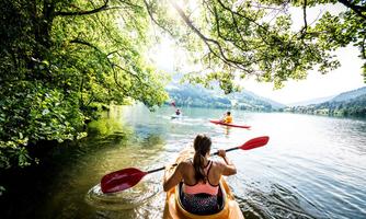 A woman is paddling in a kayak on a calm lake, surrounded by lush greenery. In the background, other kayakers can be seen, and the sun is shining pleasantly.