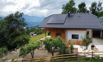 A modern house with solar panels on the roof, surrounded by trees and a lush green hill. In the background, mountains and a clear sky are visible.