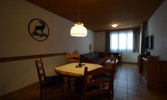 A cozy living space with a round table and wooden chairs. There is a sofa by the window and a television on the wall.