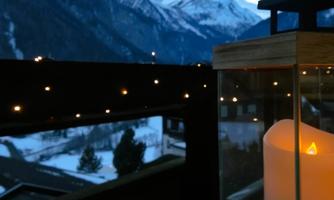 A lantern with a burning candle stands on a table. In the background, snow-covered mountains and a clear evening sky can be seen.