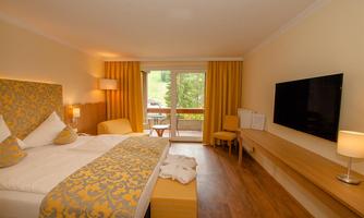 A cozy hotel room with a large bed, yellow curtains, and a TV on the wall. The room has a nice balcony overlooking nature.