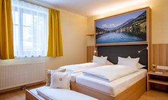 A cozy bedroom with two single beds and a large window. A picture of a picturesque lake hangs on the wall.