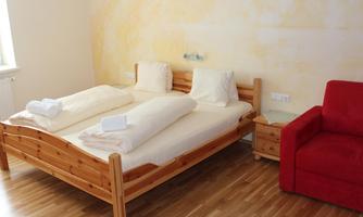 A cozy bedroom with a wooden double bed and beige sheets. Next to the bed is a red armchair and there is a light, neutral wall paint.