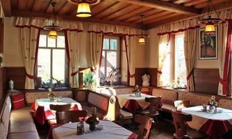 A cozy restaurant with wooden furniture and traditional decoration. The tables are covered with red tablecloths and large windows let in plenty of light.
