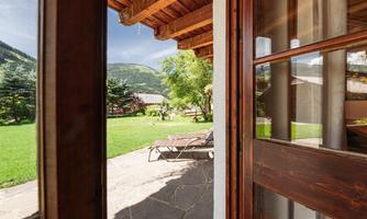A cozy view from a wooden window overlooking a green garden and gentle hills. The shutters are open and invite relaxation.