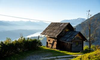 A simple, rustic wooden house stands on a hill overlooking the valley and the mountains. The landscape is picturesque and nature is peaceful.