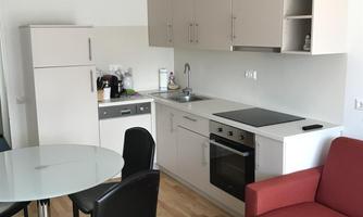 A modern kitchen with a dining table and chairs. The room is bright and friendly with wooden flooring and a red sofa.