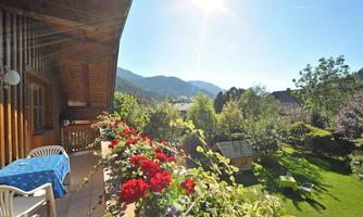 A balcony with a blue table and blooming red flowers. In the background, green trees and mountains can be seen under a sunny sky.