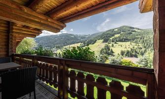 A picturesque view from a balcony overlooking green hills and forests. The clear sky and the rustic wooden structure create a relaxed atmosphere.