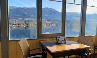A beautiful view of a lake and the surrounding mountains through large windows. A table with chairs is in the foreground.