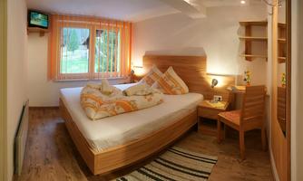 A cozy bedroom with a large bed, colorful pillows, and wooden shelves. There is a window with curtains and a small table with a chair.