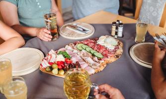 A table with a large selection of sausage and cheese platters, complemented by fresh vegetables and olives. Drinks in glasses are also on the table.