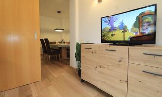 A cozy living space with wooden flooring and a television displaying a colorful animation. In the background, a dining area with a table and chairs is visible.