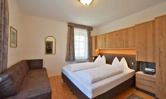 A cozy bedroom with a double bed and a couch. The walls are painted in light colors, and there is plenty of natural light.
