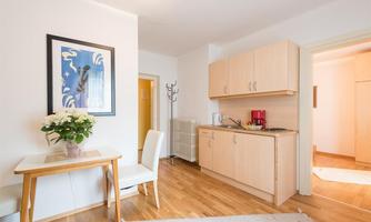 A cozy room with a wooden floor and a small kitchenette. A vase of flowers is on the table, and a picture hangs on the wall.
