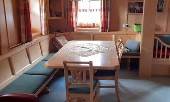 A cozy living space with a wooden table and chairs. The walls are in warm tones, and there are large windows with curtains.