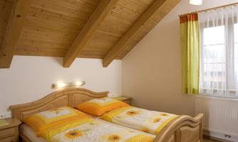 A cozy bedroom with a wooden ceiling and a double bed. The bedding is light and floral, and there is plenty of daylight through the window with curtains.
