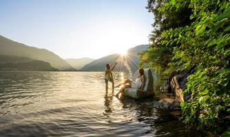 Two people are standing at the edge of a lake and enjoying the sunset. The surrounding area is surrounded by mountains and green trees.