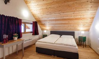 A cozy bedroom with a double bed and a wood-paneled ceiling. The walls are light and there are windows with curtains as well as a small table and nightstands.