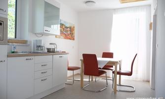 A bright, modern kitchen with a dining area. The table is equipped with white chairs and red seat cushions.