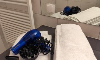 A hairdryer lies next to stacked white towels on a dark surface. In the background, a bathroom is reflected with a wall and a light switch.