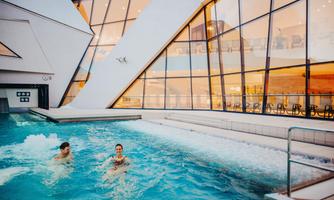 A modern indoor pool with smooth water and large glass windows. Two people are swimming relaxed in the water.