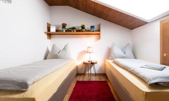 A cozy room with two single beds and a small table. On the wall, there is a shelf with plants and decorations.