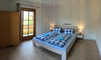 A bright bedroom with a double bed and checked bed covers. The room has large windows and a wooden wardrobe.