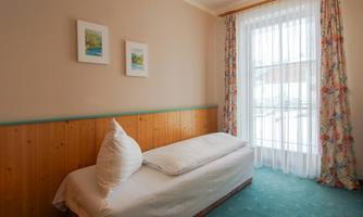 A simple room with a single bed and a large window. Bright curtains let in a lot of light and there are some decorative pictures on the wall.
