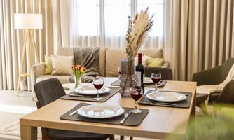 An elegant dining area with a wooden table, set with white plates and red wine. In the background, a cozy living room can be seen with a sofa and large windows.