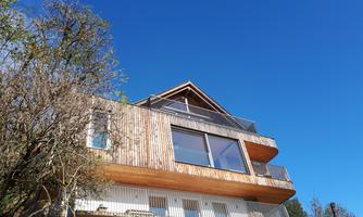 A modern wooden house with large windows and a balcony. The sky is clear and blue, surrounded by trees.