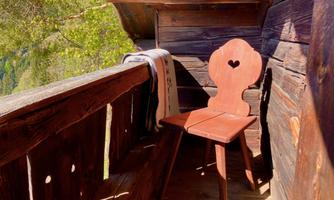 A beautiful wooden chair with a heart-shaped backrest stands on a balcony. In the background, green trees and warm sunlight can be seen.