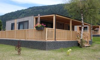 A modern holiday home with a wooden porch in a green environment. In the background, gentle hills and trees can be seen.
