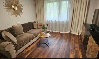A cozy living room with a beige sofa and a small table. A window with curtains lets in a lot of daylight.