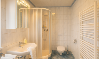 A modern bathroom with a shower, a sink, and a toilet. The walls are light tiled and there is a towel warmer.
