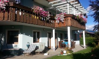 A beautiful house with blooming balcony plants and a cozy garden. There are loungers and an outdoor dining area.