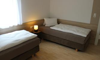 A simply furnished room with two single beds and a nightstand. The window lets in plenty of light, and there is soft lighting.