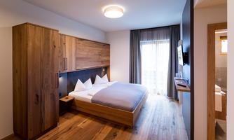 A modern bedroom with wooden furniture and a large bed. The windows allow plenty of natural light to come in and provide a view of a balcony.