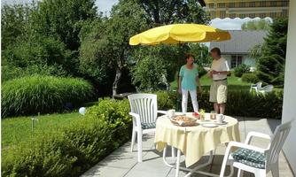 A beautiful terrace with a yellow sun umbrella and a table set for breakfast. In the background, there is a green garden with trees and bushes.