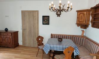 A cozy dining area with a wooden table and rustic chairs. The walls are light and the decor is traditional, featuring a beautiful chandelier and a towel on the table.