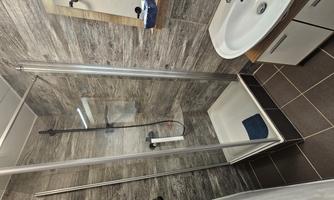 A modern bathroom with a glass shower enclosure and a sink. The walls are designed in a wood look and the tiles are dark.