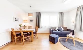 A bright and modern living room with a gray sofa and a wooden dining table. Large windows provide plenty of daylight and a cozy atmosphere.