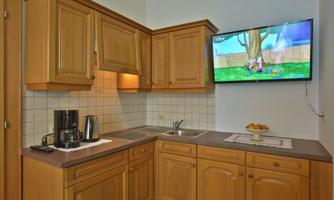 A cozy kitchen with wooden furniture and modern appliances. A television is mounted on the wall, showing a colorful cartoon series.