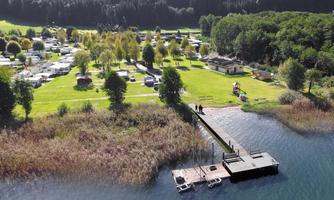 A beautiful campsite by the water with a green meadow and a pier. In the background, trees and RVs can be seen.