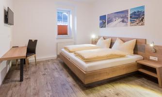 A modern bedroom with a double bed and wooden furniture. Landscape paintings hang on the wall, and a small table and a chair are available.