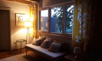 A cozy living room with a bright couch and cushions. Large windows let the evening light in, while the yellow curtains create a warm atmosphere.