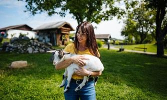 A woman is holding a small goat in her arms. In the background, green areas and a rural building can be seen.