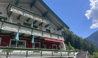 A traditional multi-story building with a red and white facade paint. There are balconies with green railings and blue awnings, surrounded by mountains and trees.