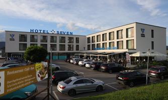 A modern hotel with the sign "Hotel Seven". Many cars are parked in front of the building, and there is a terrace with seating.
